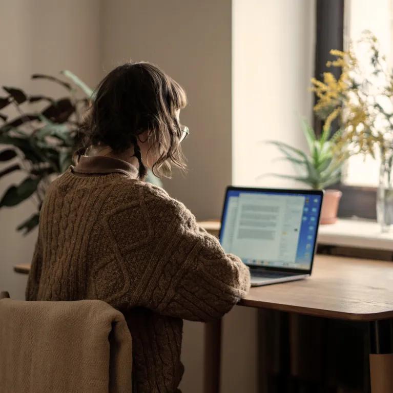 A woman wearing a brown jumper, working on a laptop at a wooden desk in a room with houseplants and natural light from a nearby window