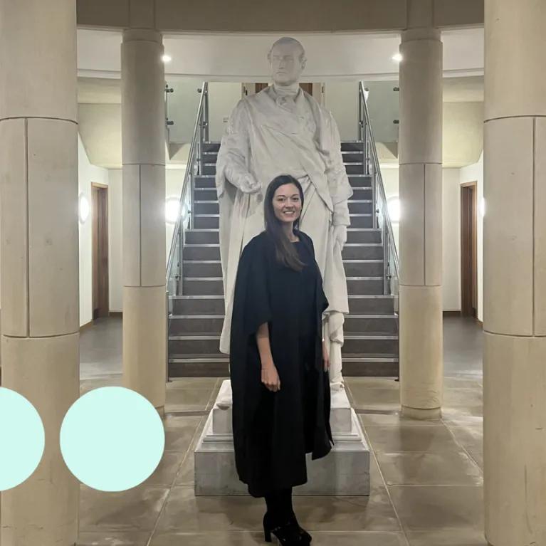 Woman in graduation gown stood in front of a statue 
