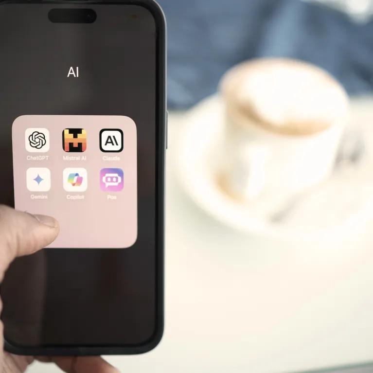 A person holding a mobile phone displaying AI-related apps including ChatGPT, with a blurred background showing a coffee on a table