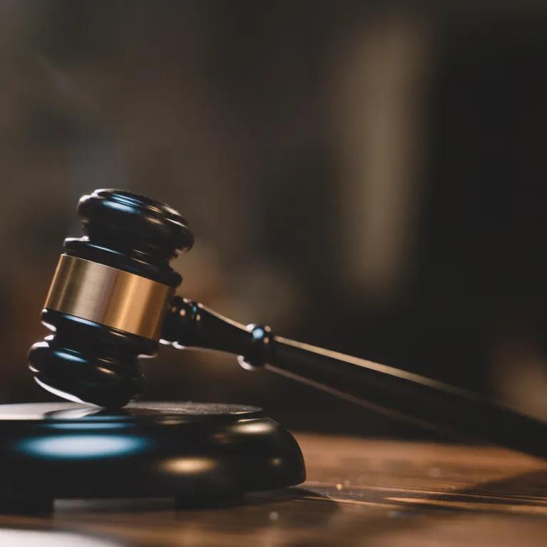 A black and gold gavel resting on its block, with the head tilted and handle touching a wooden table. The background shows a blurred courtroom