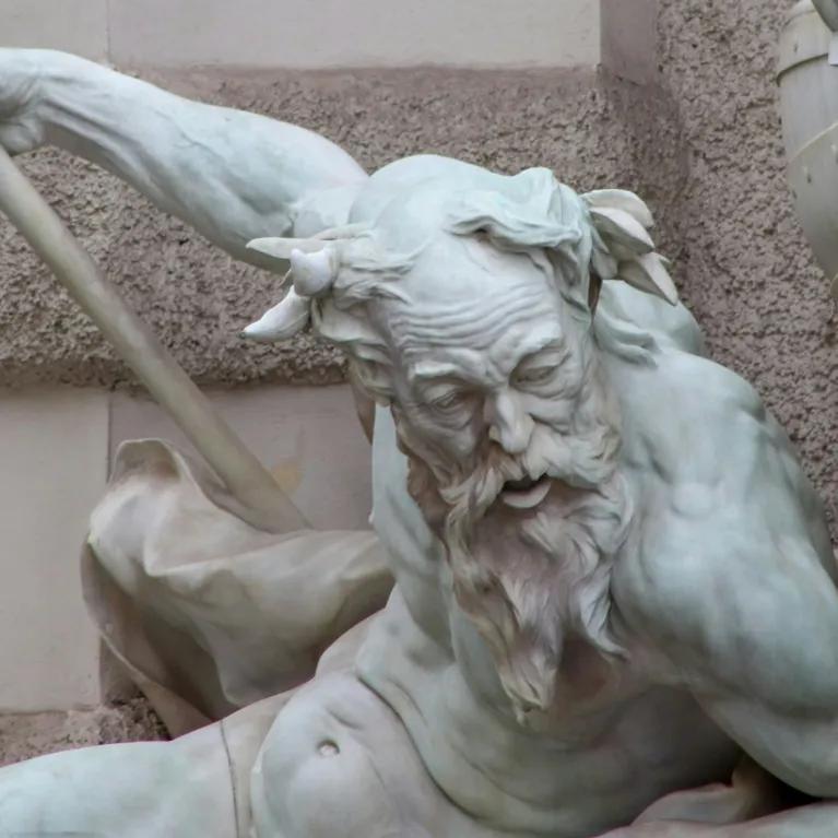 A marble statue of a muscular, bearded man with horns, gripping a staff and leaning forward in a dynamic pose. Another figure and a large shield are partially visible beside him, suggesting a mythological battle scene.