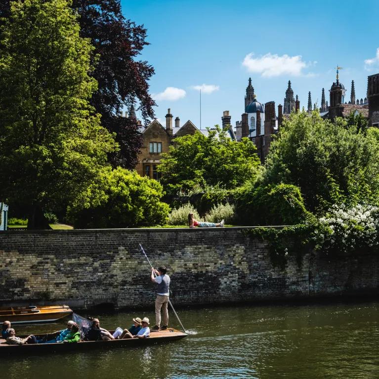 A punt full of people goes along the river cam against the backdrop of a college garden full of trees and greenery.