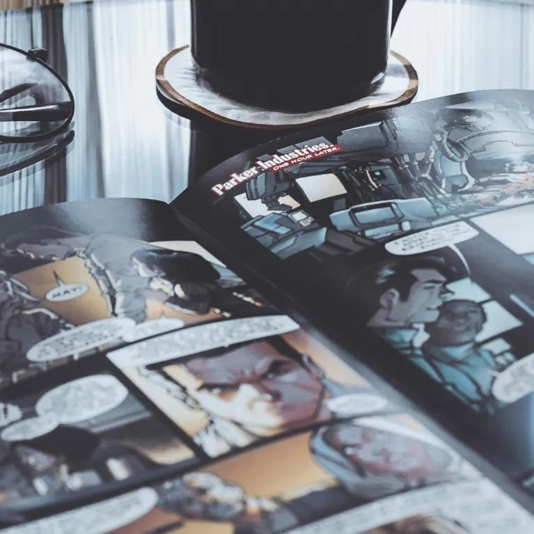 An open comic book lying on a glass table, with a black mug of coffee beside it and a pair of round glasses resting on the comic’s pages.