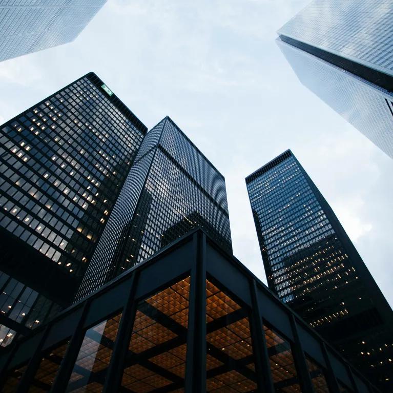 A low-angle view of several lit-up skyscrapers against a cloudy evening sky.