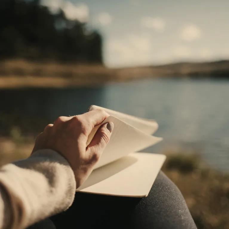 A person’s hand turning a page in a notebook while sitting by a lake, with blurred trees and sky in the background