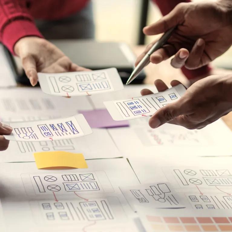 Several hands pointing to a mind map covered in post-it notes during a brainstorming session, with more notes and leaflets being held