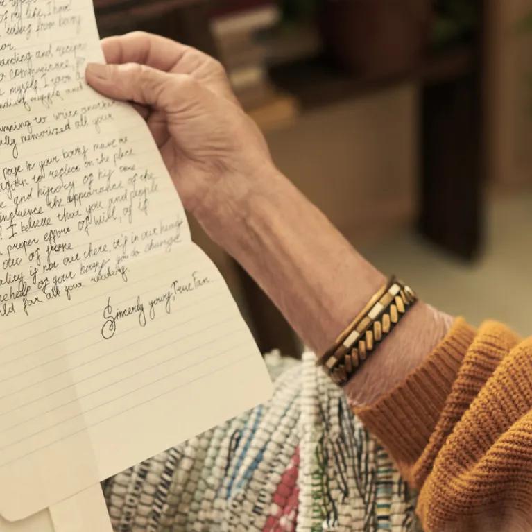 An elderly woman with glasses and a gold bracelet on her right wrist, holding and reading a handwritten letter, with the focus on the letter and her hands.