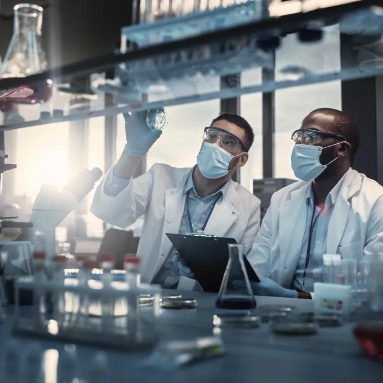 Two scientists wearing masks, lab goggles, and white coats examining a cell culture plate in a laboratory, conical flasks and test tubes are blurred in the background.