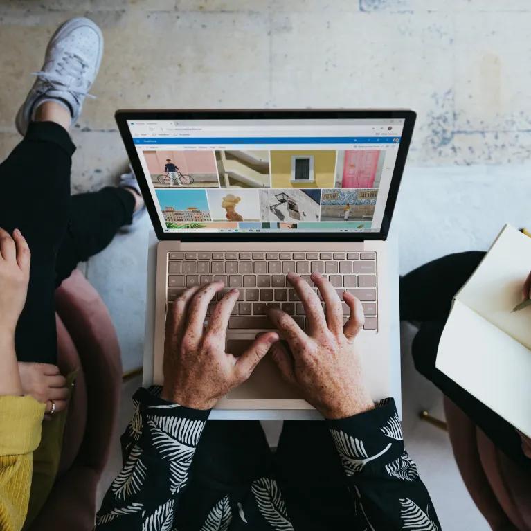 Overhead shot of three people working on a laptop and taking notes, with a Microsoft Surface Laptop displaying a photo collage on the screen.