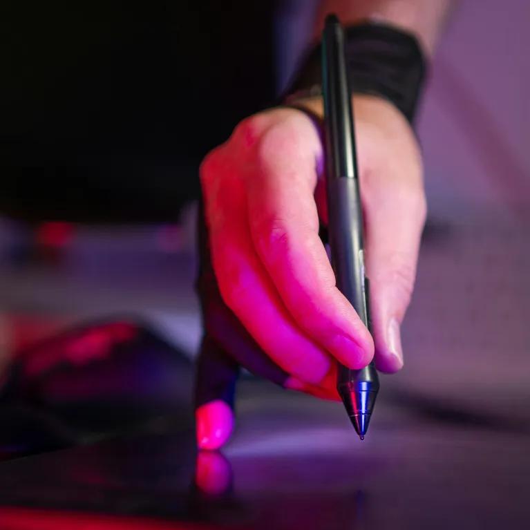 A person's hand, illuminated by red light, holds a digital pen over a drawing tablet, with a black wristband visible on their wrist and a dark background. 