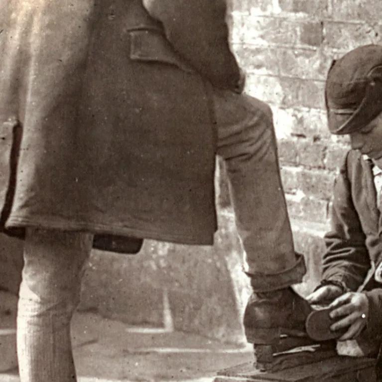 A black and white photo of a boy shining the shoes of a person. 