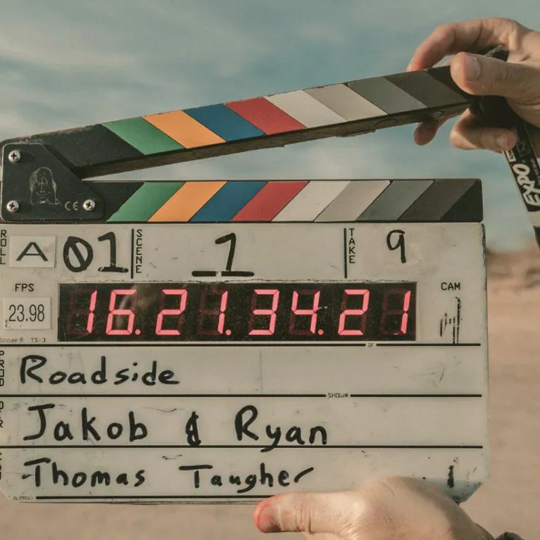 A close-up shot shows a film clapperboard being held by two hands against a blurred background of a desert landscape under an overcast sky.