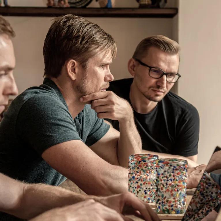 Three men are gathered around a laptop on a wooden table.