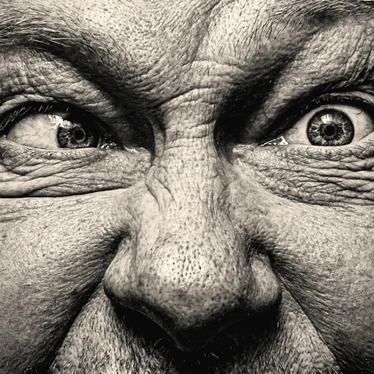 Close-up, high-contrast black and white portrait of a man with intense, wide eyes and wrinkled forehead, looking to the side with one eye and forward with the other.