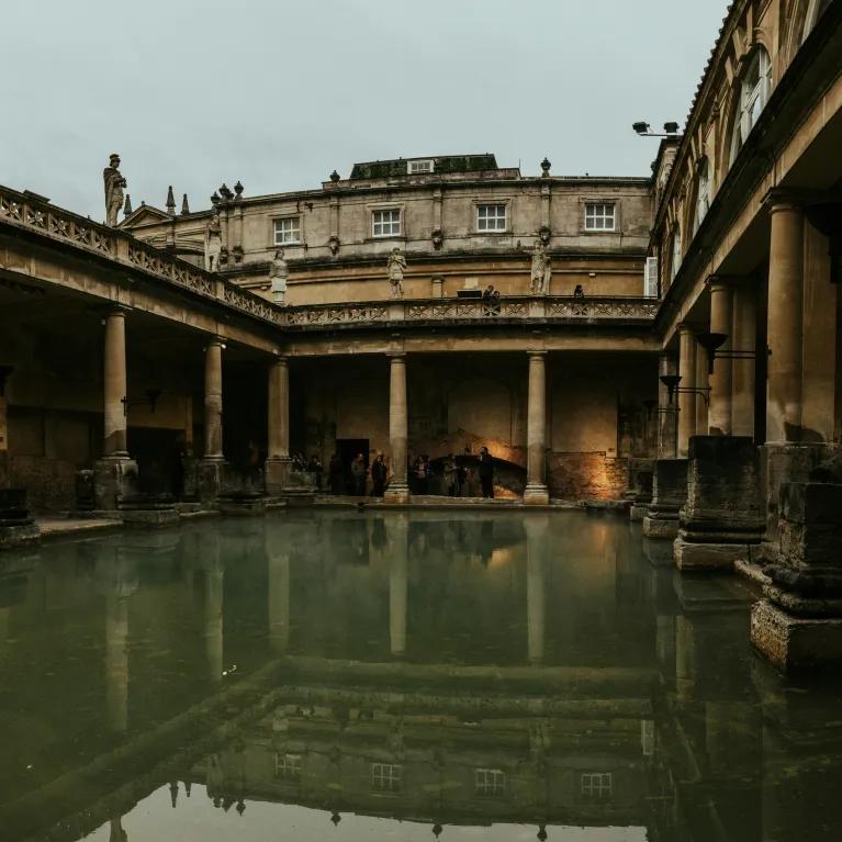 A wide shot of the Great Bath at the Roman Baths , featuring the large, green-tinged water of the main pool surrounded by ancient Roman architecture, including large columns and statues on the upper terrace, under an overcast sky."