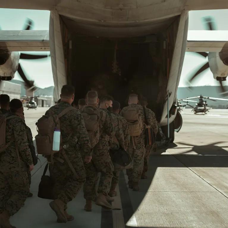 A group of military personnel, identified as US Navy SEALs, are boarding a large military transport aircraft, likely a C-130 Hercules, on an airfield with other helicopters visible in the background. 