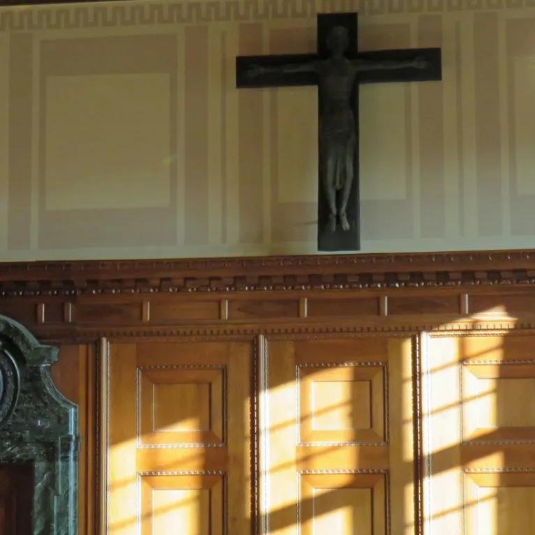 A room with wooden paneling on the lower walls and lighter-colored walls above, featuring two ornate crystal chandeliers and a large, dark crucifix mounted centrally on the upper wall. Sunlight streams through a window, casting shadows on the wooden panels.