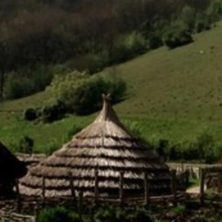 A painting of rustic wooden huts with thatched roofs set against green hills, trees, and dense woodland in the background, with a lone man walking away into the scene.
