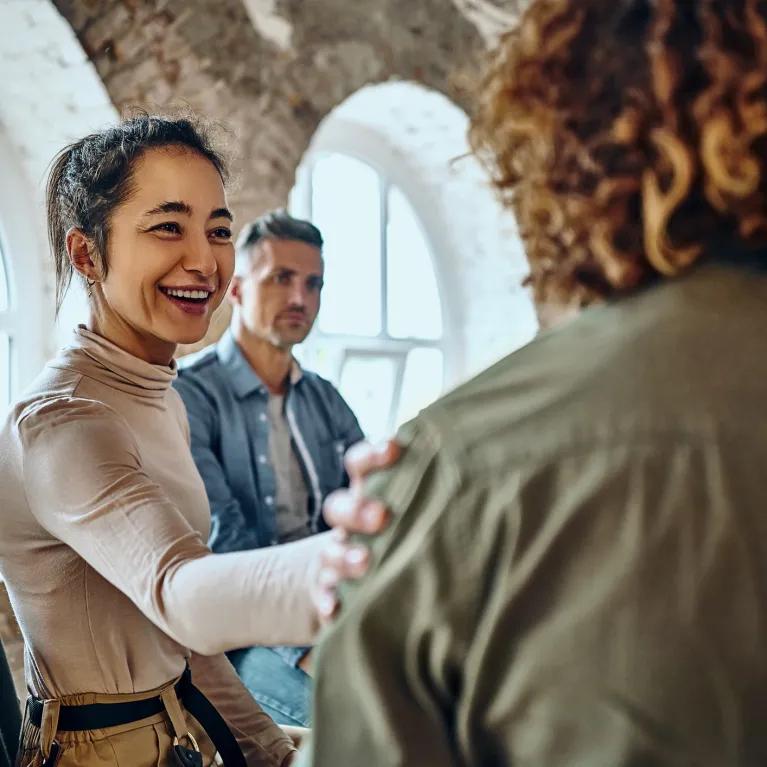 A smiling woman touches another person's shoulder in a supportive group setting, with a third person in the background.