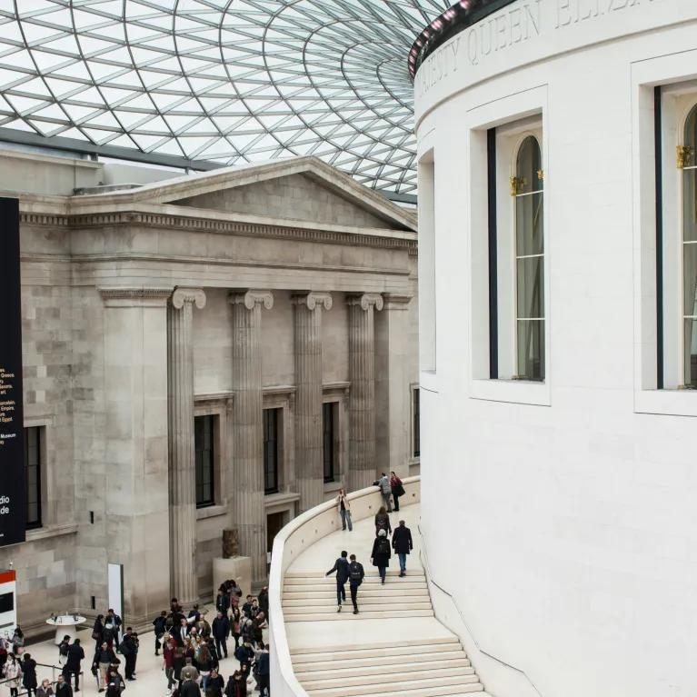 An interior shot of the British Museum's Great Court in London, featuring the vast tessellated glass roof and the circular Reading Room, with numerous visitors traversing the open space and climbing the grand staircase, visible under daylight filtering through the roof.