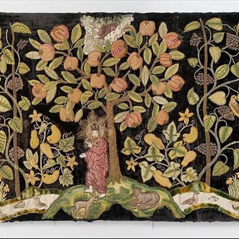 A late 16th-century British embroidered textile depicting 'The Garden of Eden' on dark velvet, featuring vibrant green trees laden with colorful fruits, flowers, and leaves, with figures of Adam and Eve amidst the lush landscape and various animals.