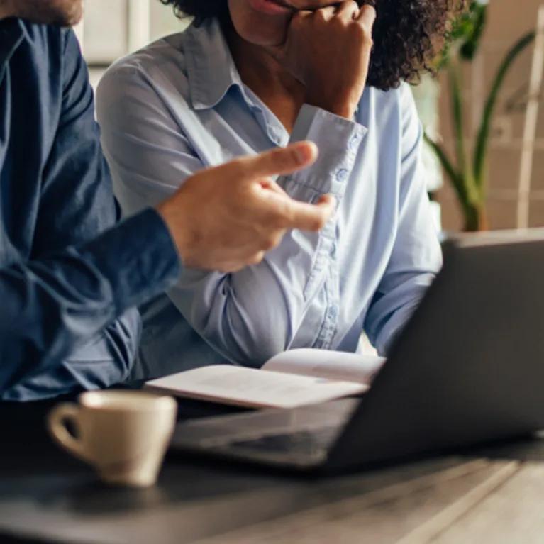 A man and a woman sitting together at an open laptop on a table, having a conversation.