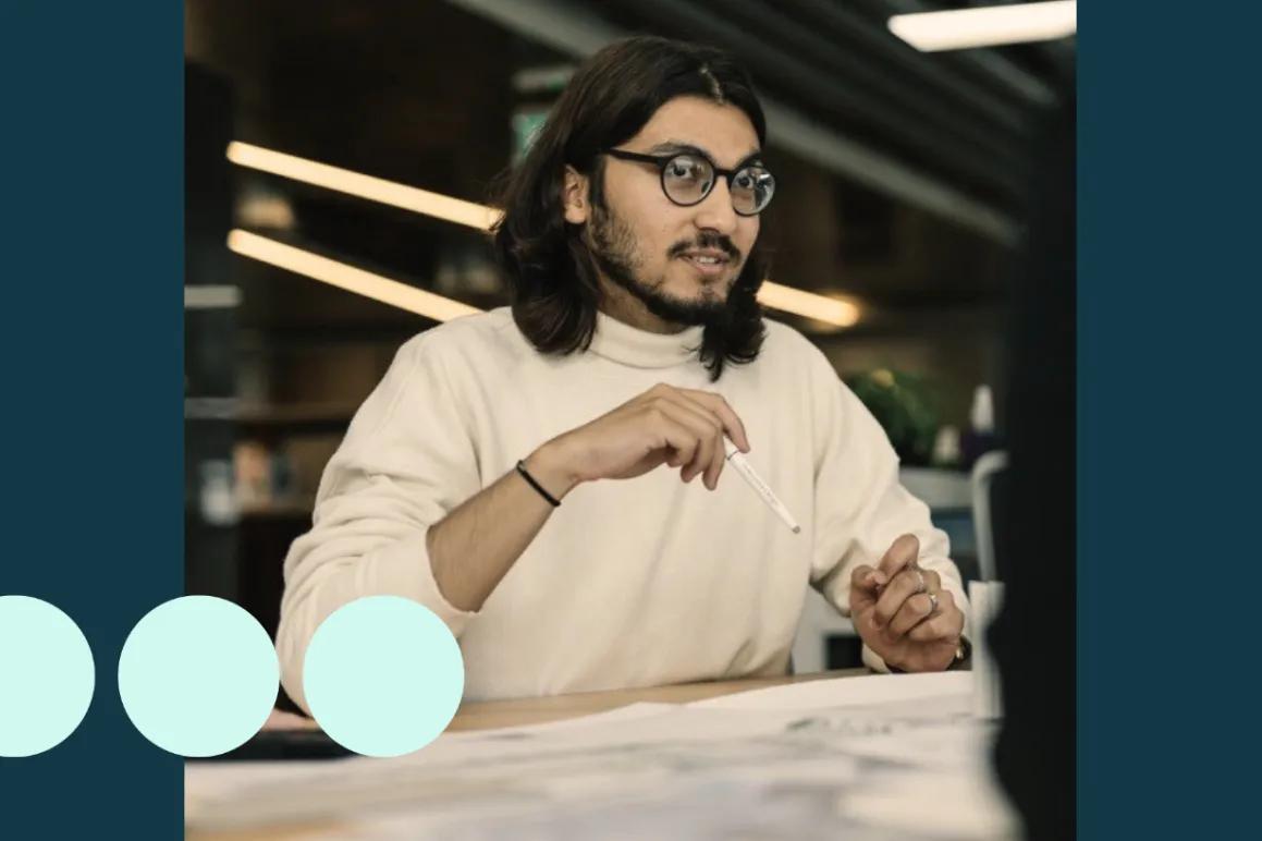 Man with shoulder length dark hair, sat at desk with pencil in hand