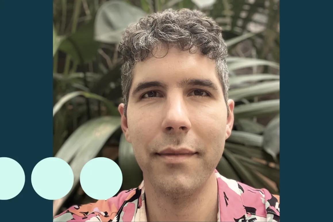 Man with greying dark curly hair, stood in front of large plant 